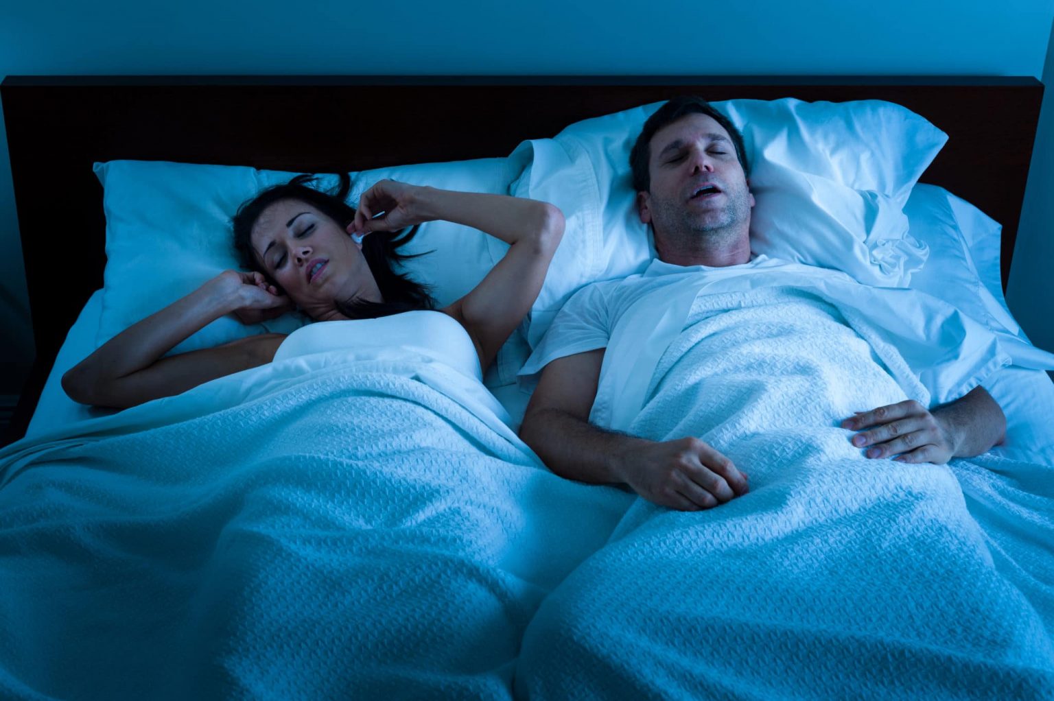 snoring-solutions Man and woman in bed and the woman is covering her ears due to the man snoring.